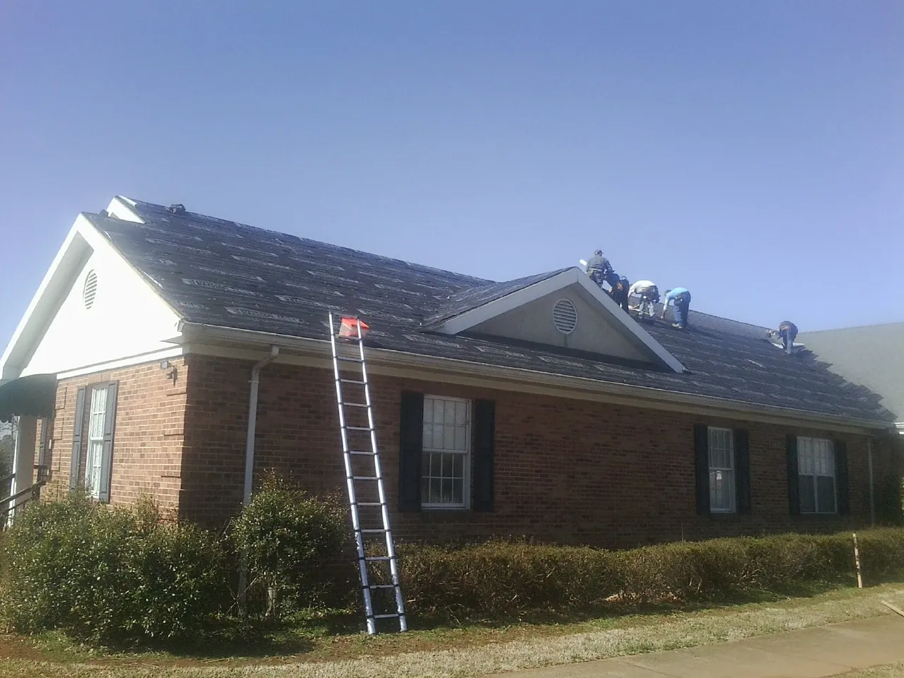 Crew tearing shingles off a brick ranch roof with a ladder set — Metal Roof Repair work by our crew in Burnt Hills