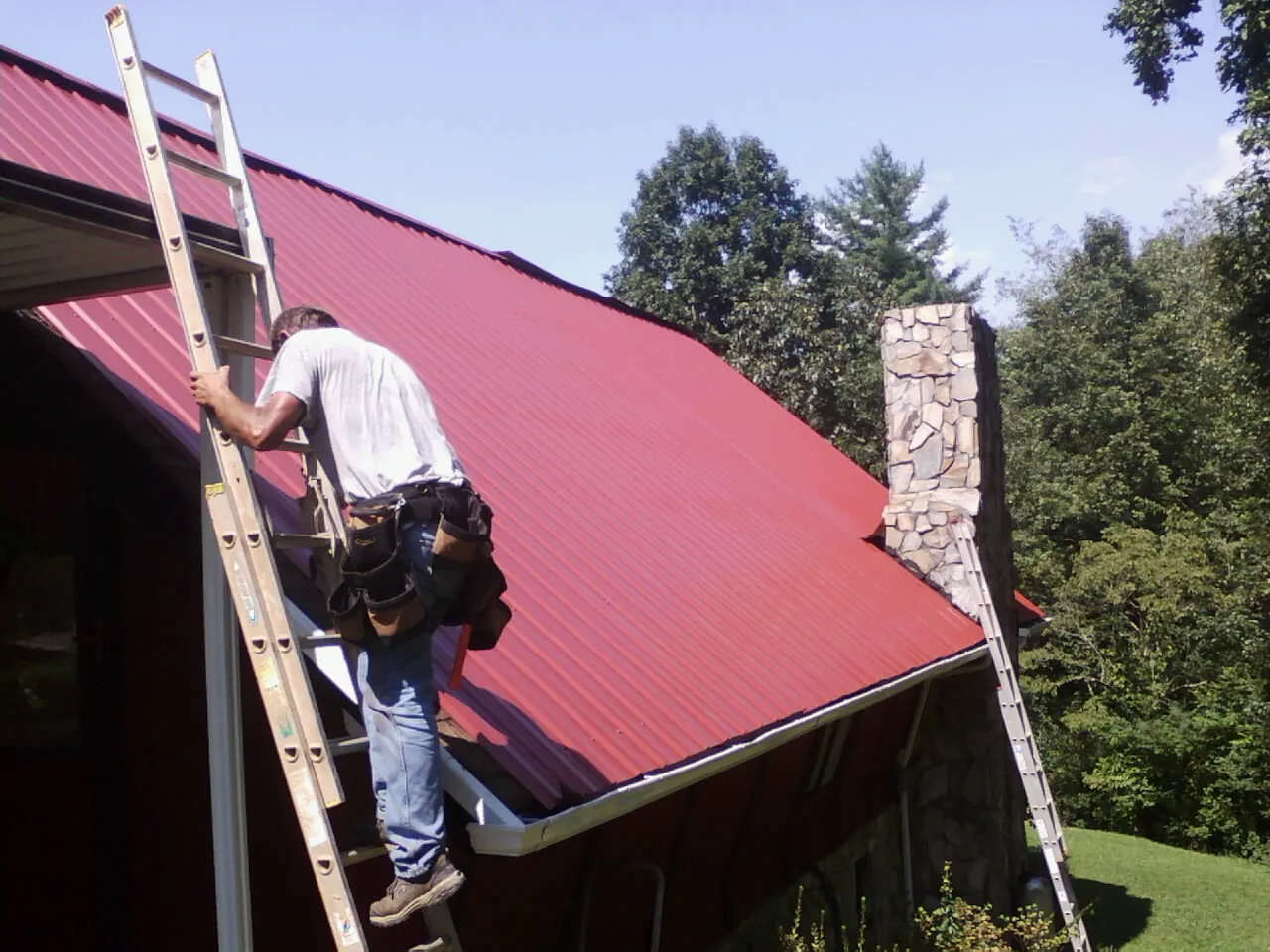 Roofer on a ladder beside a red metal roof and stone chimney — Soffit & Fascia Repair by Roofing Brothers in Burnt Hills